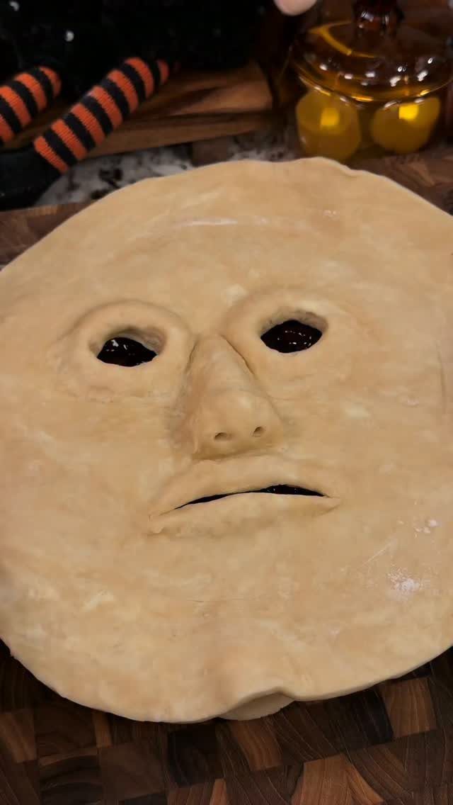 Ghost-faced berry pie with a sculpted crust and dark purple filling on wood.
