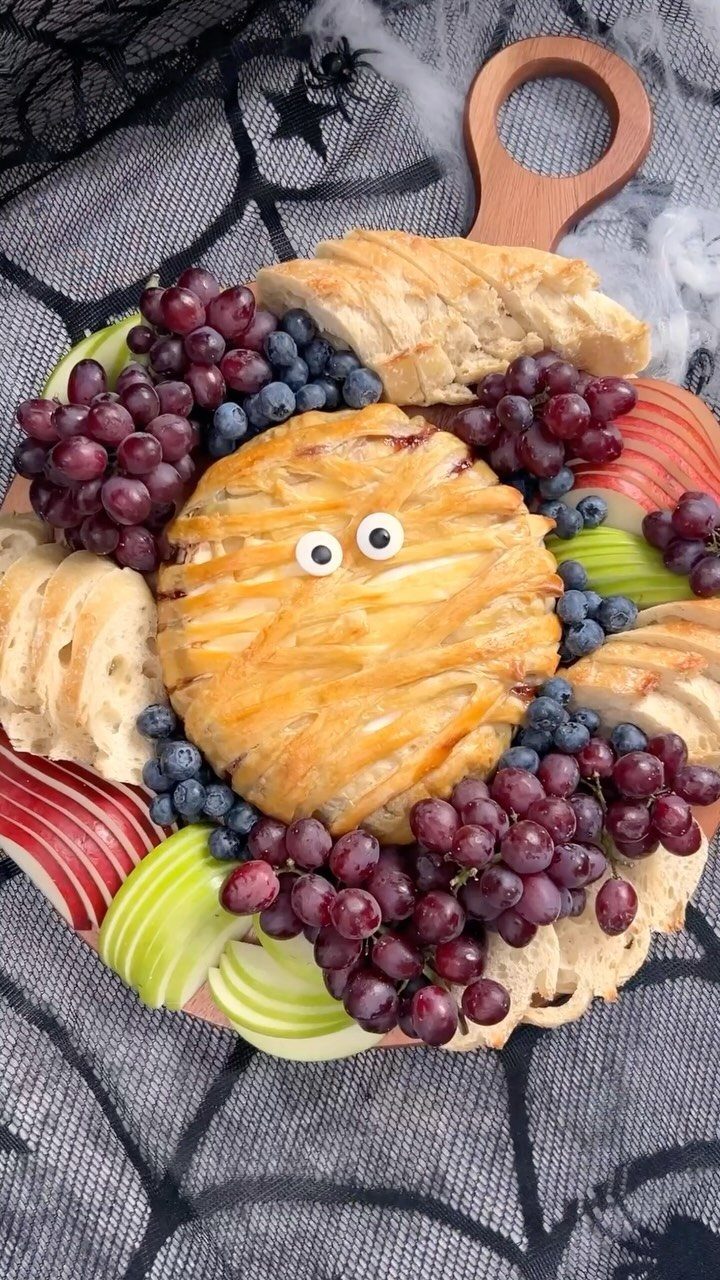 Mummy brie wrapped in pastry with candy eyes on a fruit and bread board.