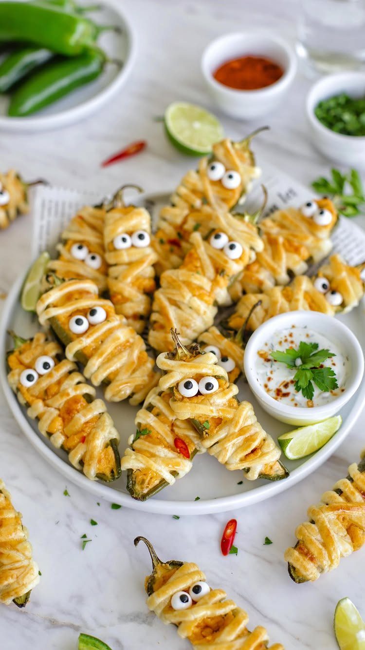 Mummy jalapeño poppers wrapped in cheese with candy eyes on a white plate.