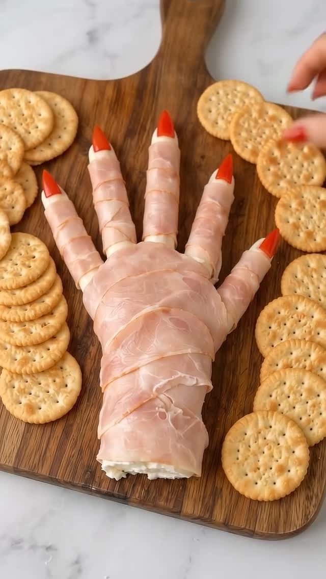 Ham-wrapped severed hand appetizer with red nails on a wooden board with crackers.