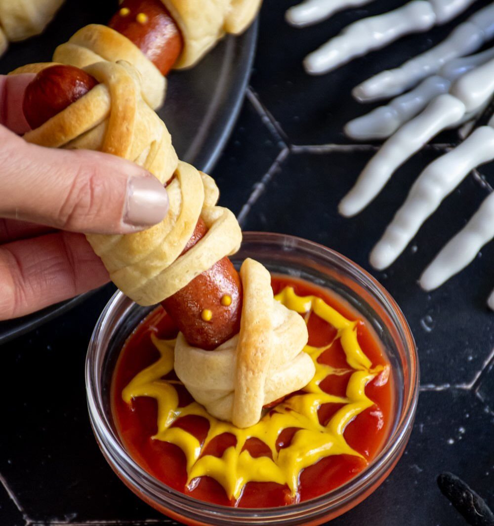 Mummy hot dog being dipped into a spiderweb mustard and ketchup sauce bowl.