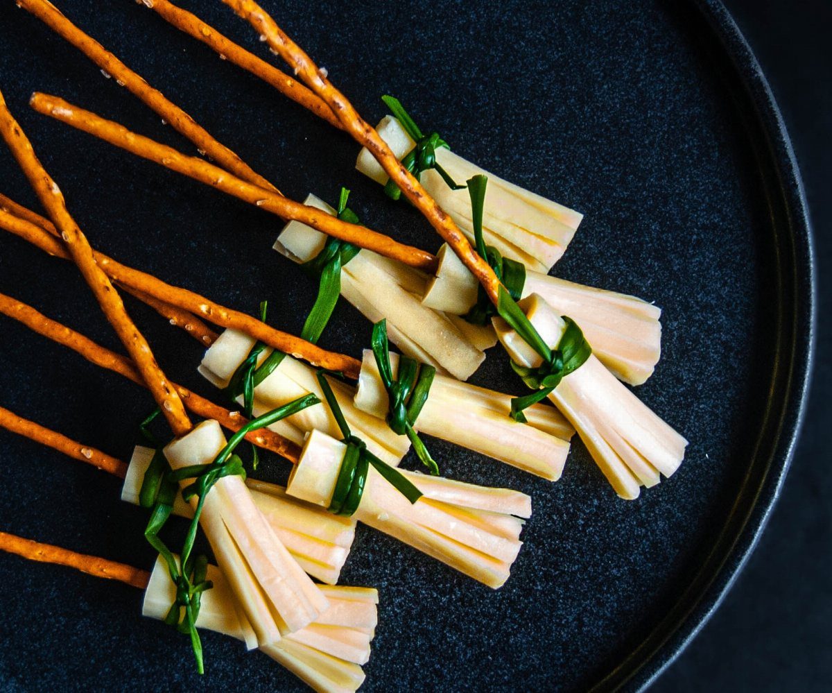 Pretzel rods wrapped in cheese strips and tied with chives on a dark plate