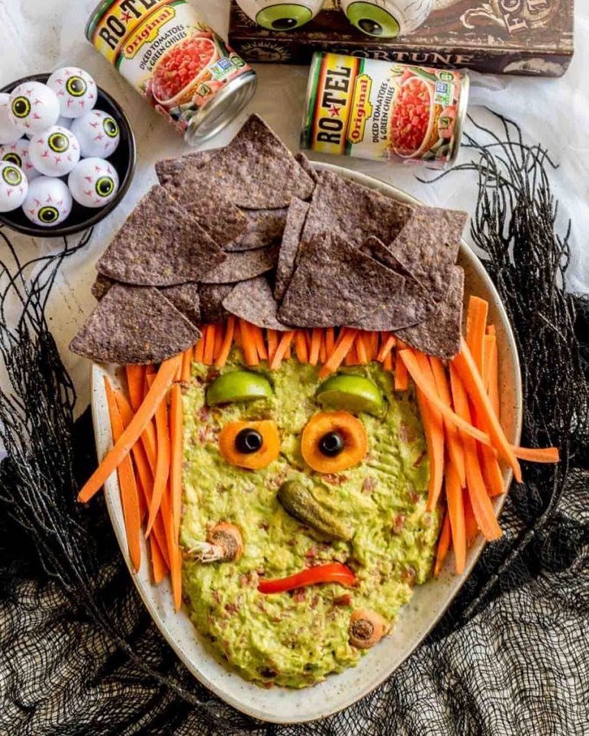 Monster face guacamole with carrot hair and blue corn tortilla chips on a platter.