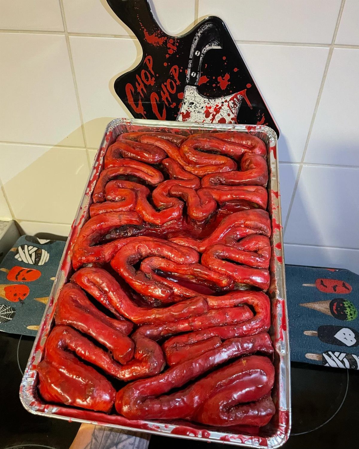 Coiled red-glazed sausages resembling intestines in a foil tray with a themed background.
