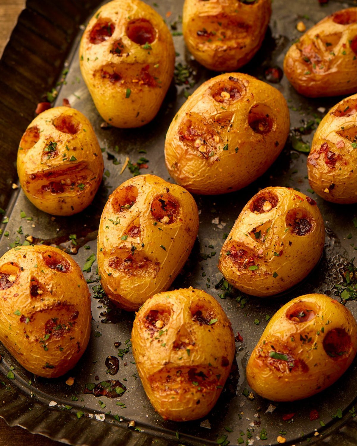 Roasted yellow potatoes carved into skull shapes on a dark baking tray.