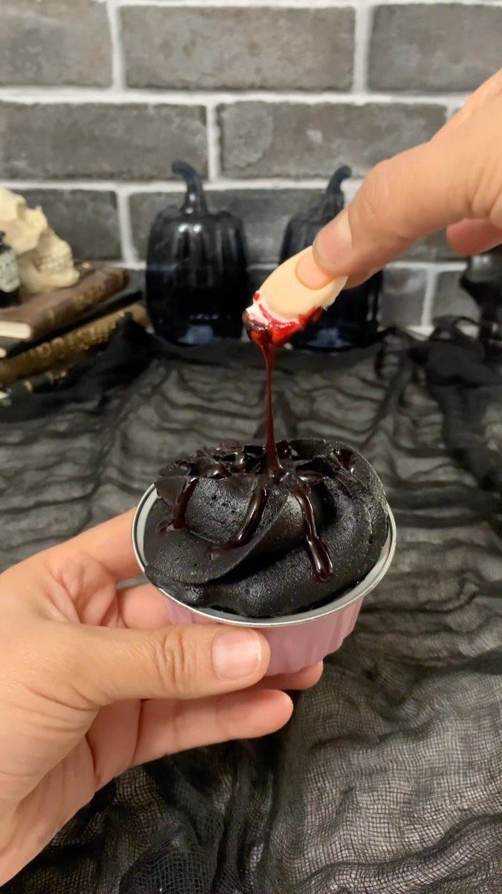 Black frosted cupcake with a finger-shaped cookie dripping red syrup being dipped in.