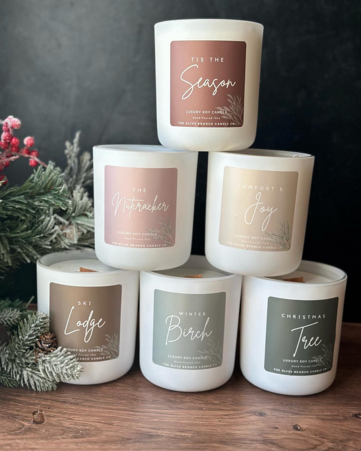 Six white holiday-themed scented candles stacked on a wooden table with pine branches.