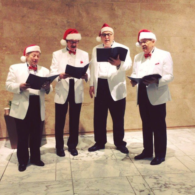 Four men in Santa hats and white blazers singing from black songbooks.