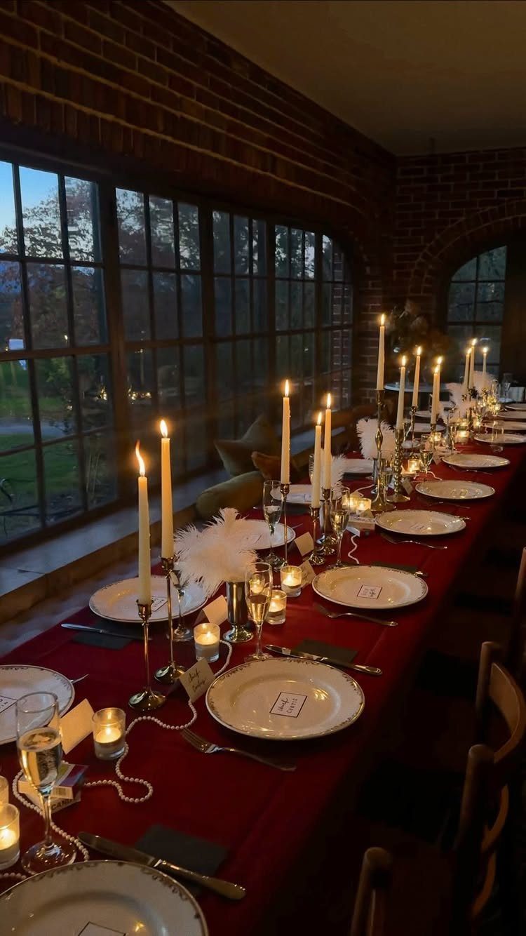 Long dining table with red cloth, white plates, and many lit taper candles.
