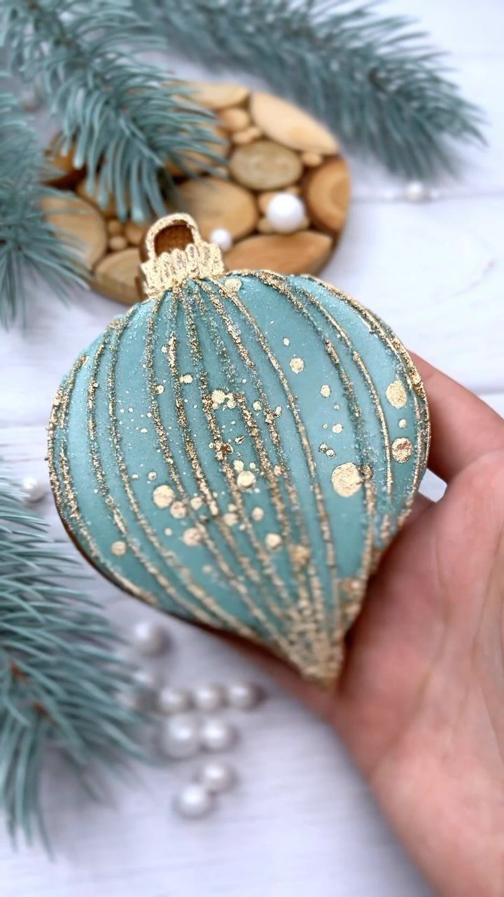 Light blue ornament-shaped cookie with gold icing held by a hand near pine needles.