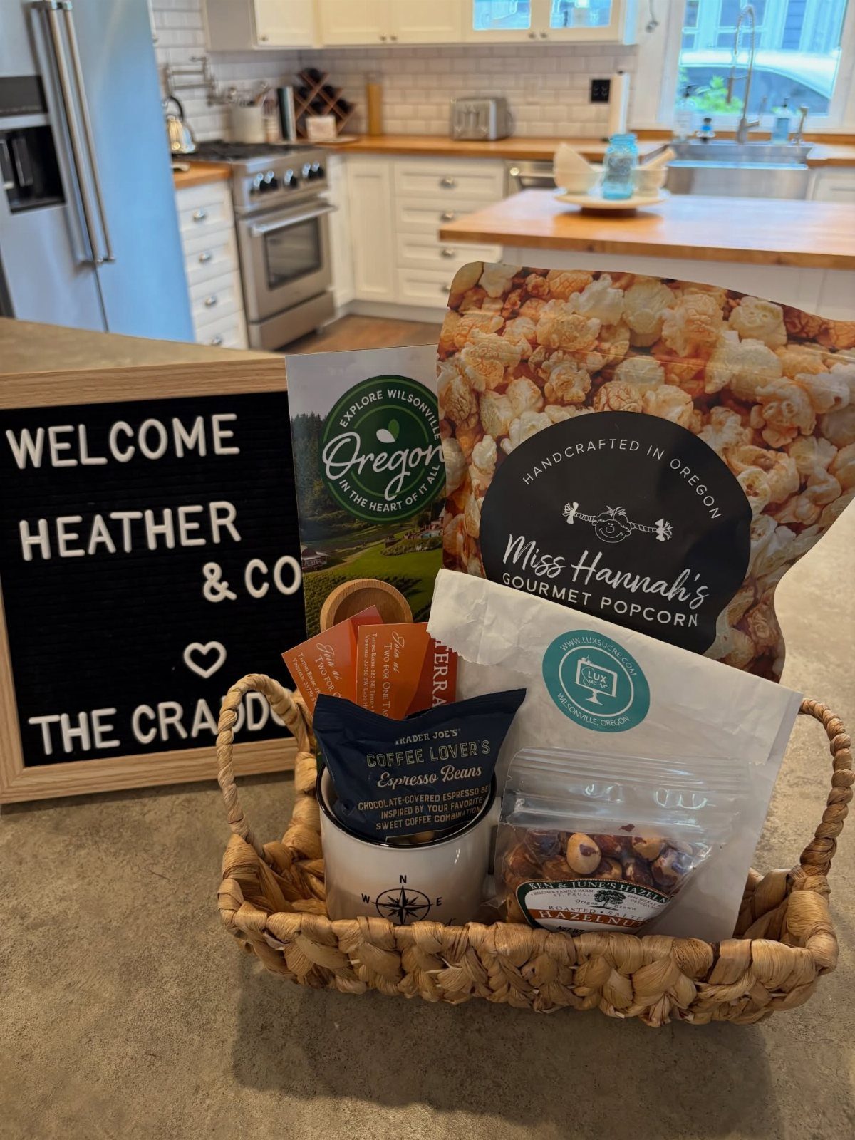 Woven welcome basket with gourmet snacks and a personalized welcome sign in a kitchen.