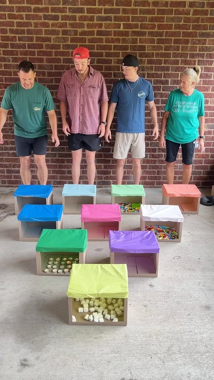 Four people standing behind colorful wooden boxes containing small objects on concrete.