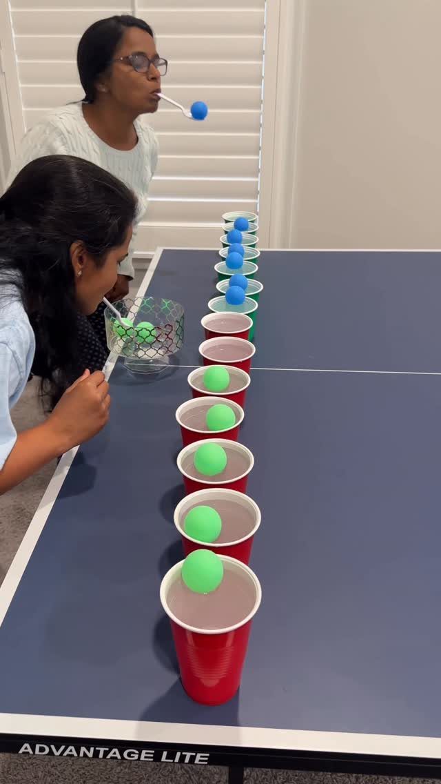 Two women blowing ping pong balls across red cups on a ping pong table