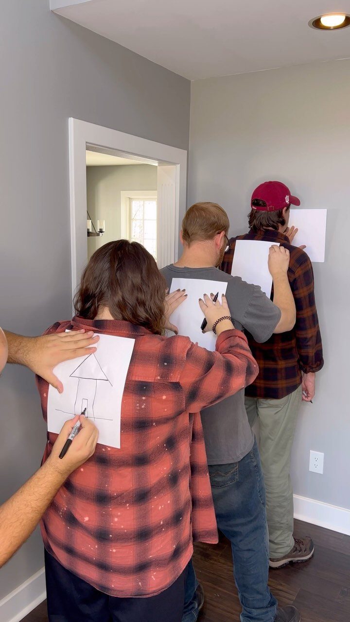People with papers on their backs, one person drawing a house on another.