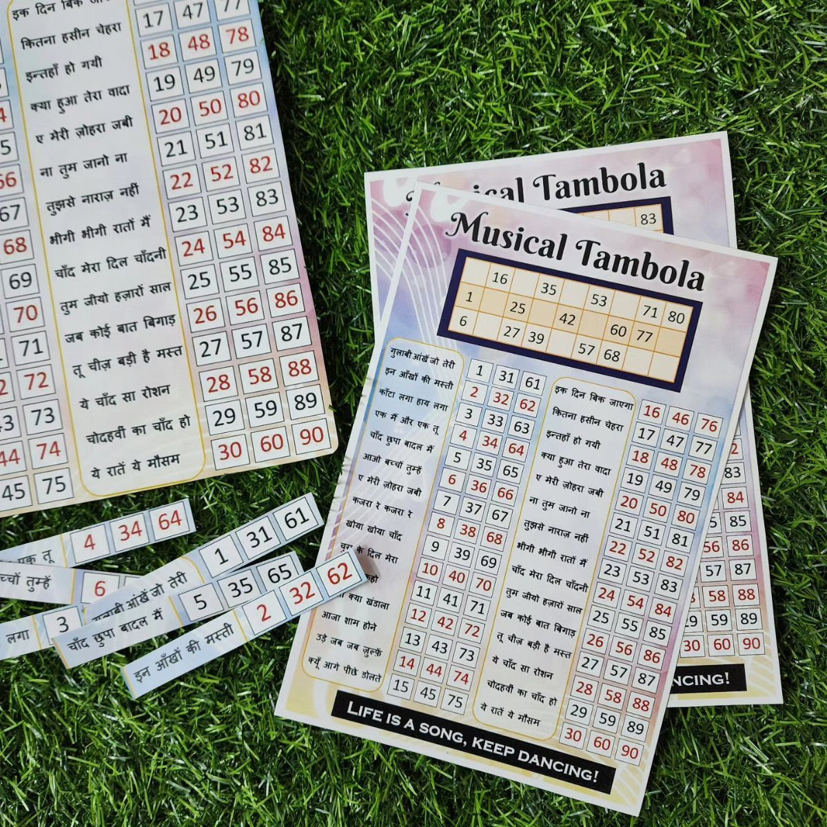 Musical Tambola game cards and number strips resting on green artificial grass.