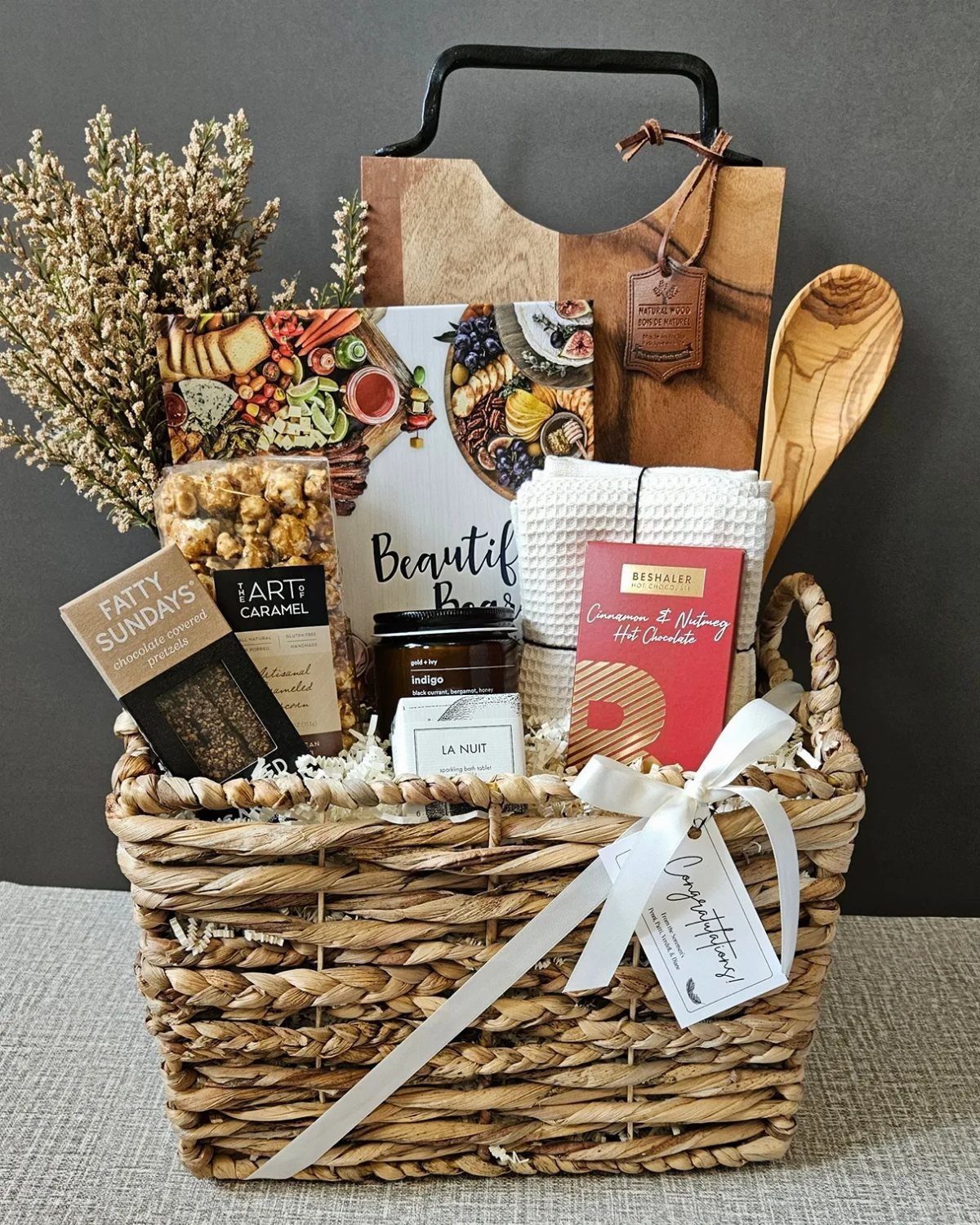 Woven gift basket containing a wooden charcuterie board, gourmet snacks, and a cookbook.