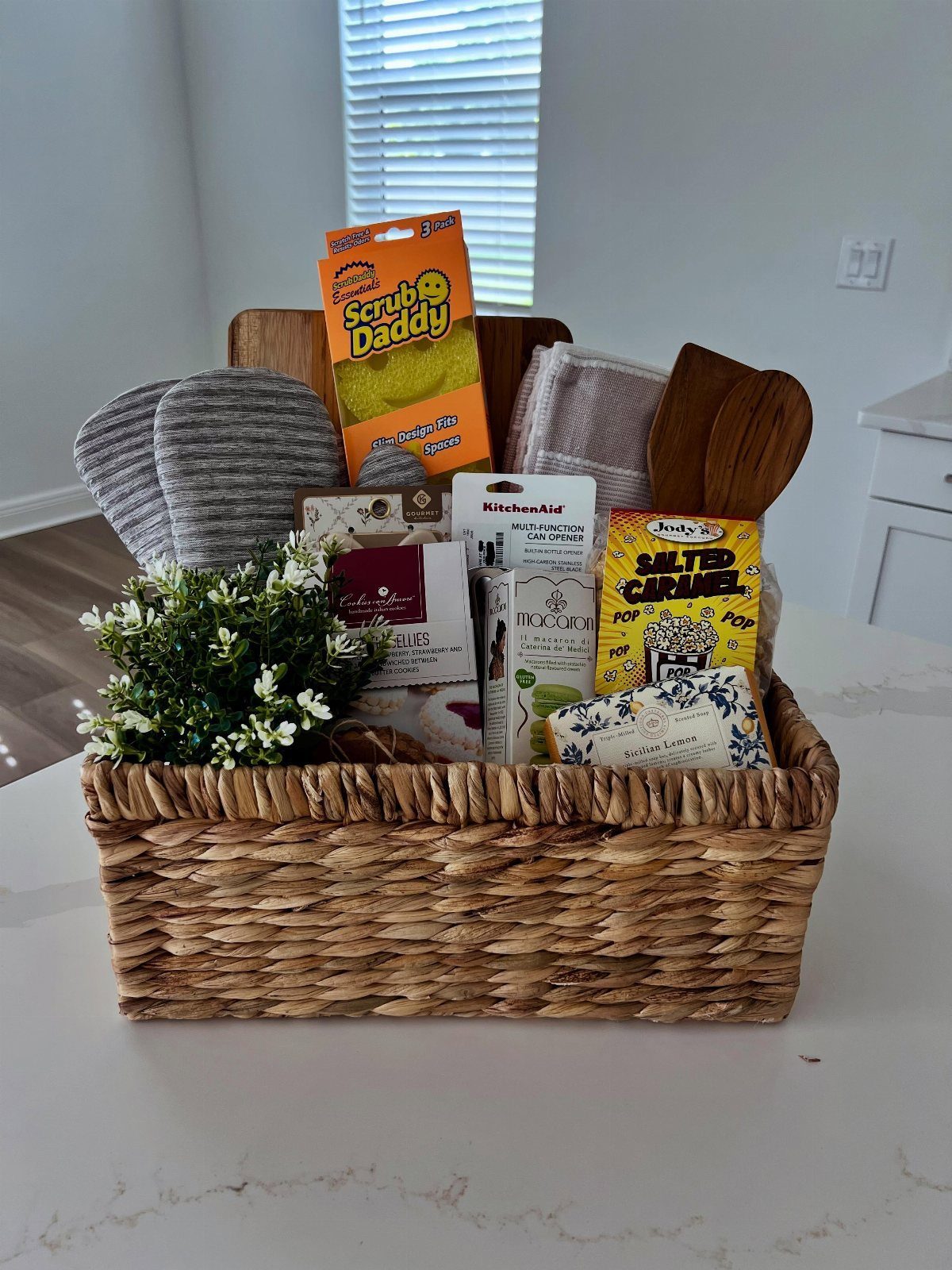 Woven gift basket containing a cleaning sponge, slippers, plant, and gourmet snacks.