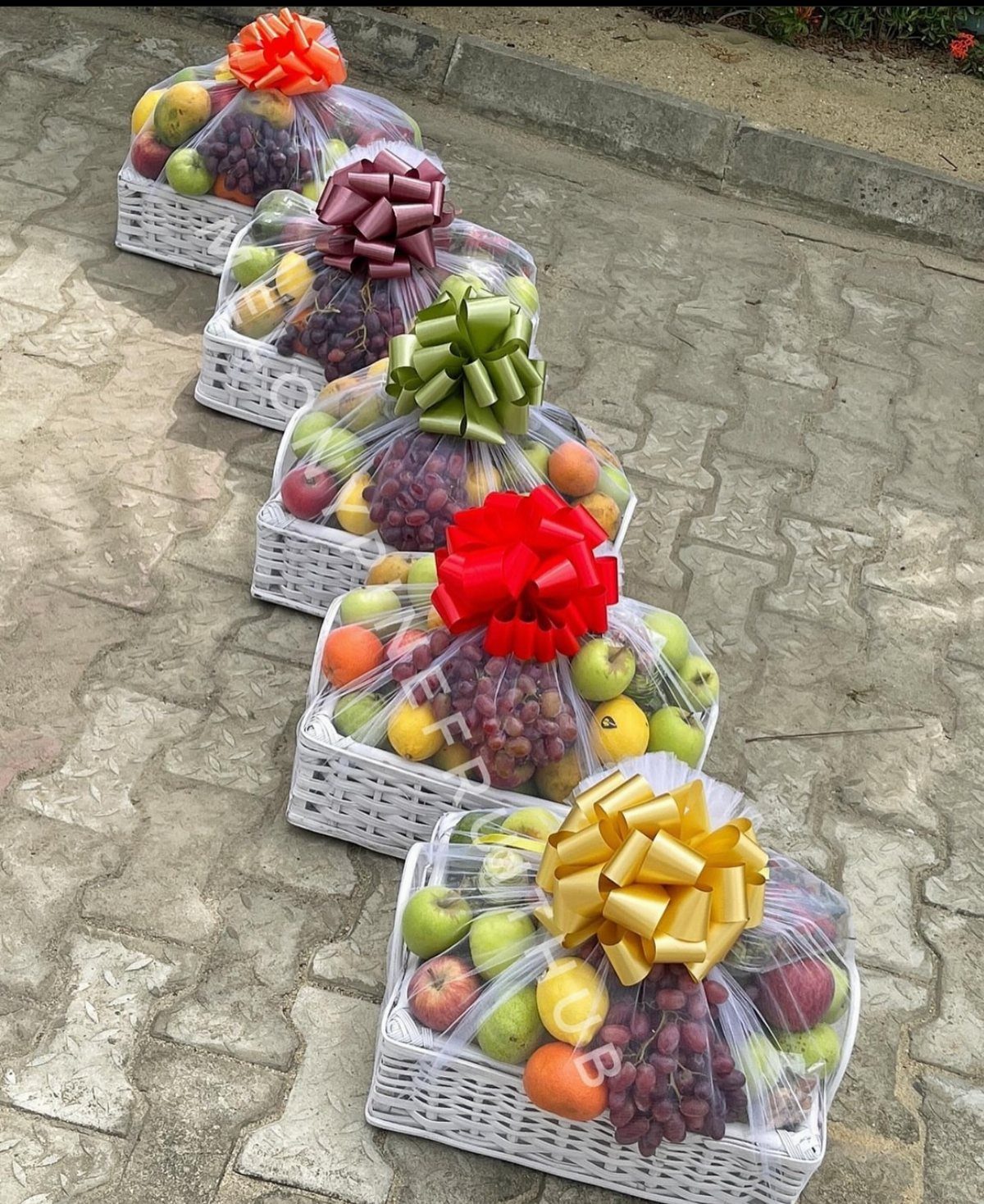 Five white wicker fruit baskets with colorful bows arranged on a stone pavement.