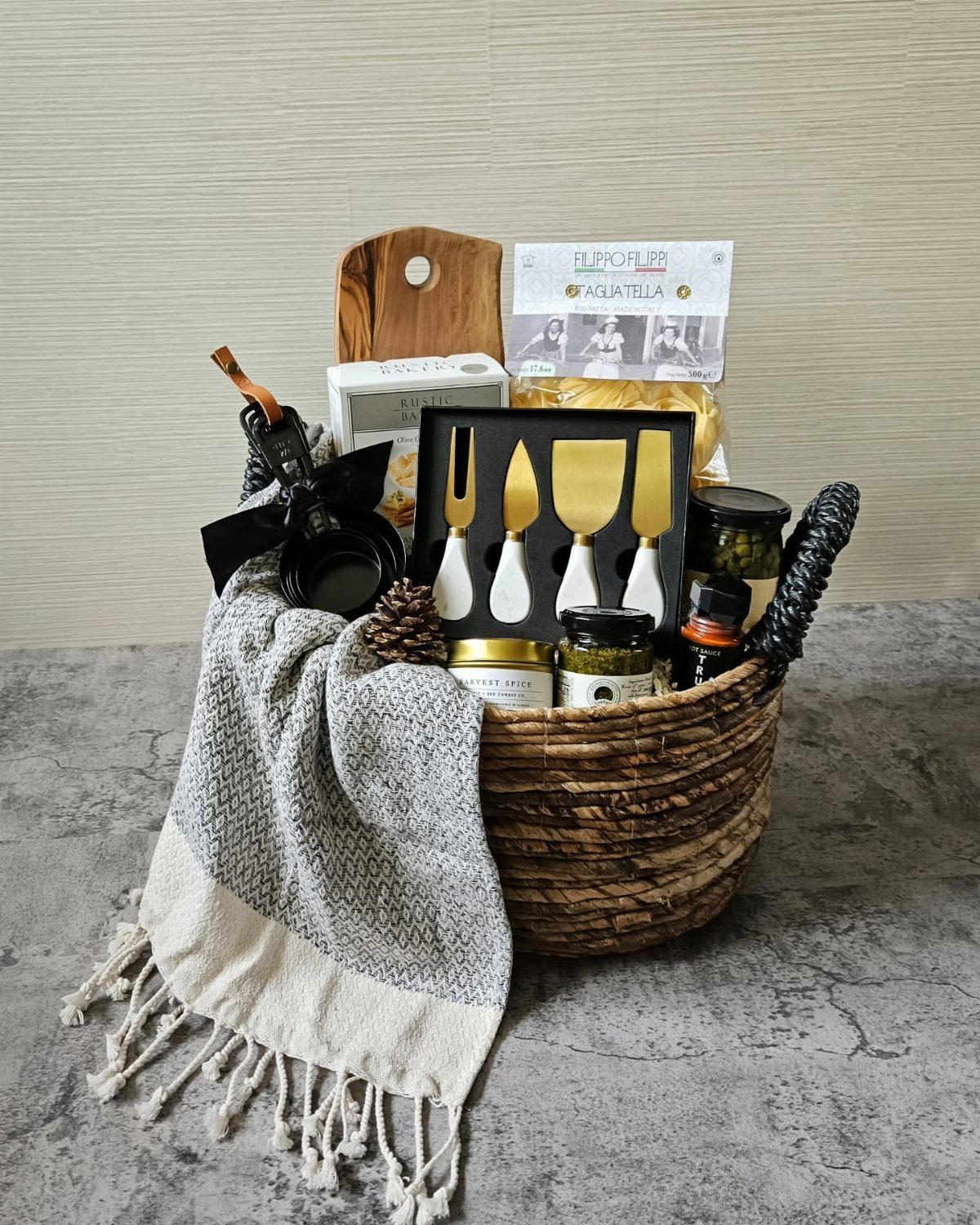 Woven gift basket containing gourmet pasta, cheese knives, and jars of preserves.