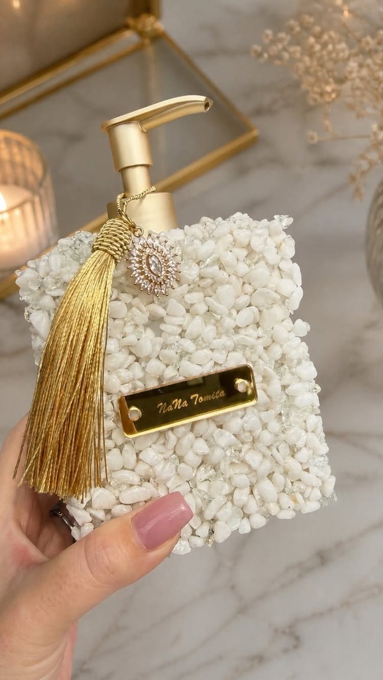 White textured soap dispenser with gold pump, tassel, and jewel held by hand.