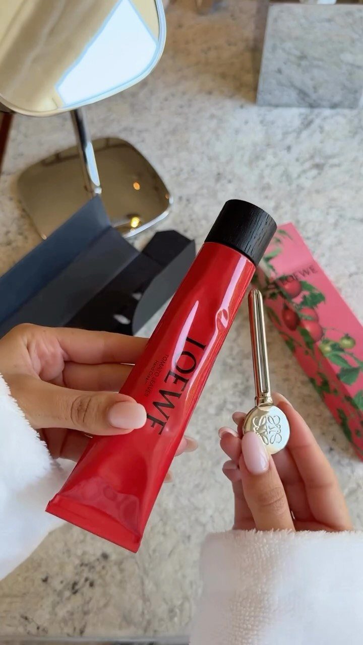 Hands holding a red tube of luxury hand cream and a gold accessory.