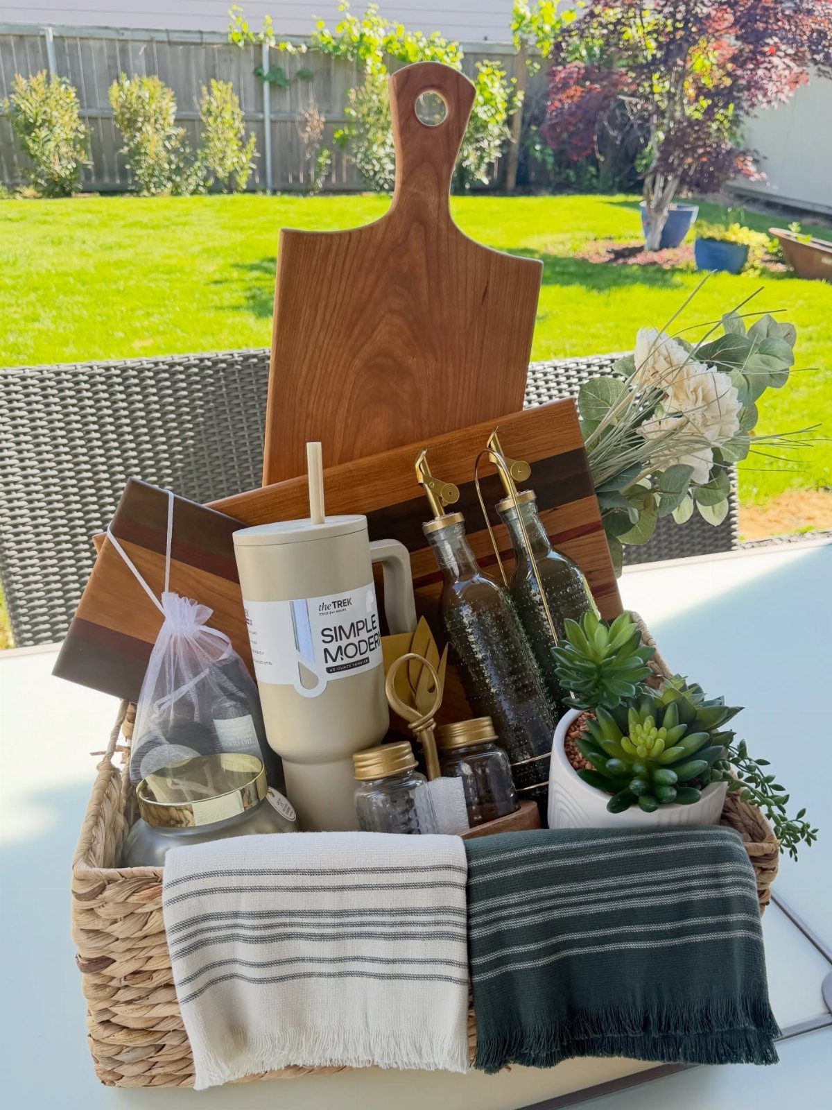 Woven gift basket with wooden cutting board, mug, bottles, and towels outdoors.