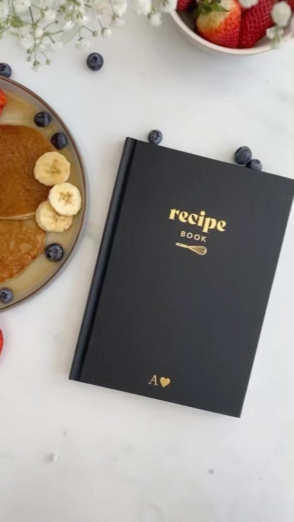 Black recipe book with gold text on a marble table with breakfast food.