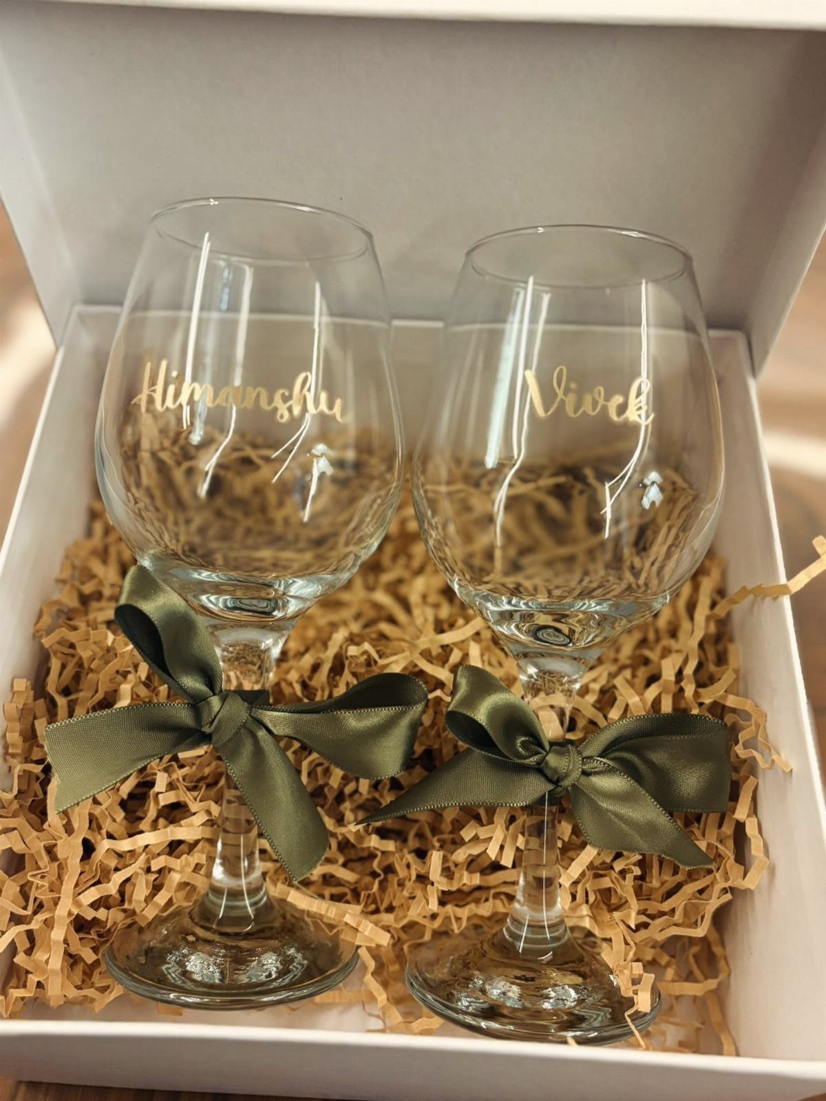 Two personalized wine glasses with gold names and green bows in a gift box