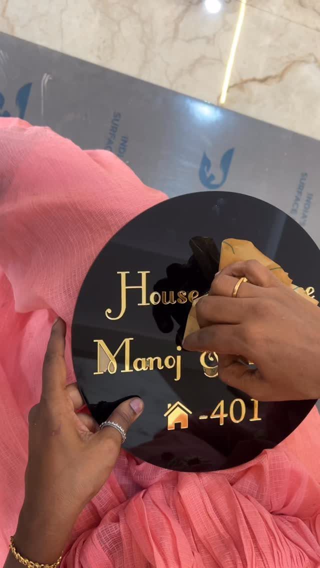 Circular black name plaque with gold lettering held by hands in pink clothing