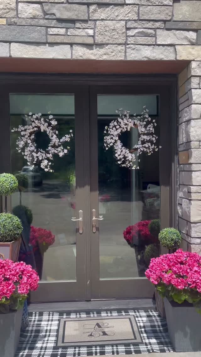 Checkered doormat and beige welcome mat in front of grey double doors with wreaths.