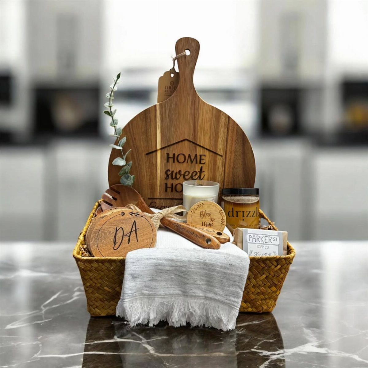 Woven wicker gift basket containing a wooden cutting board and kitchen accessories on marble.
