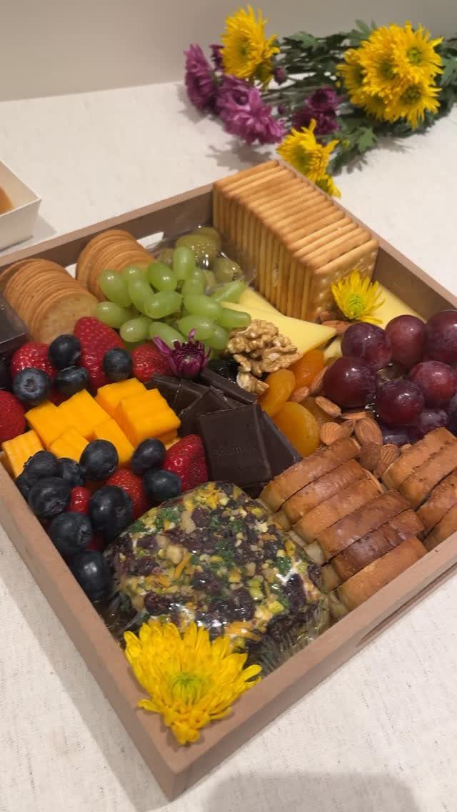 Charcuterie board in a wooden tray with fruits, cheeses, crackers, and yellow flowers.