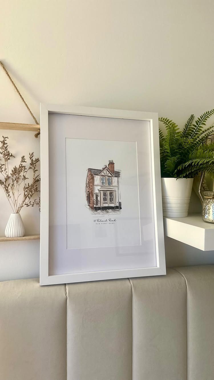 Framed custom house portrait leaning on a beige headboard next to a fern.