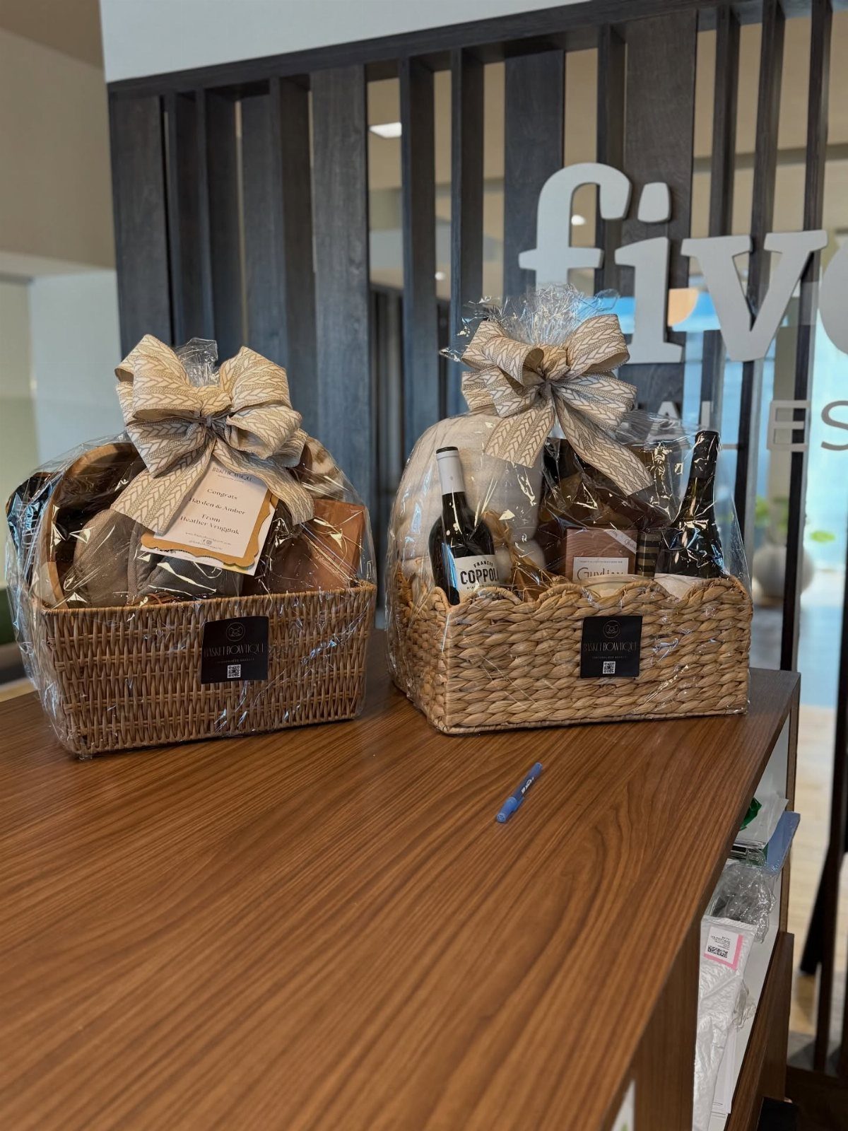 Two wicker gift baskets with wine bottles and gourmet treats on a wooden counter.