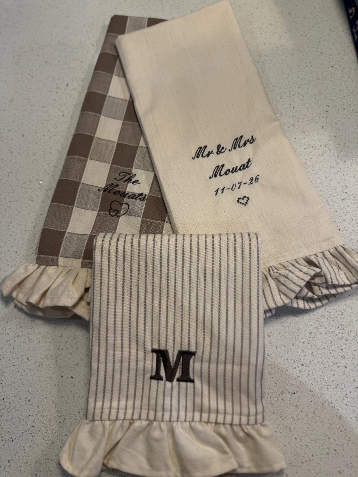 Three personalized kitchen towels with ruffles and embroidery in neutral tones on white.