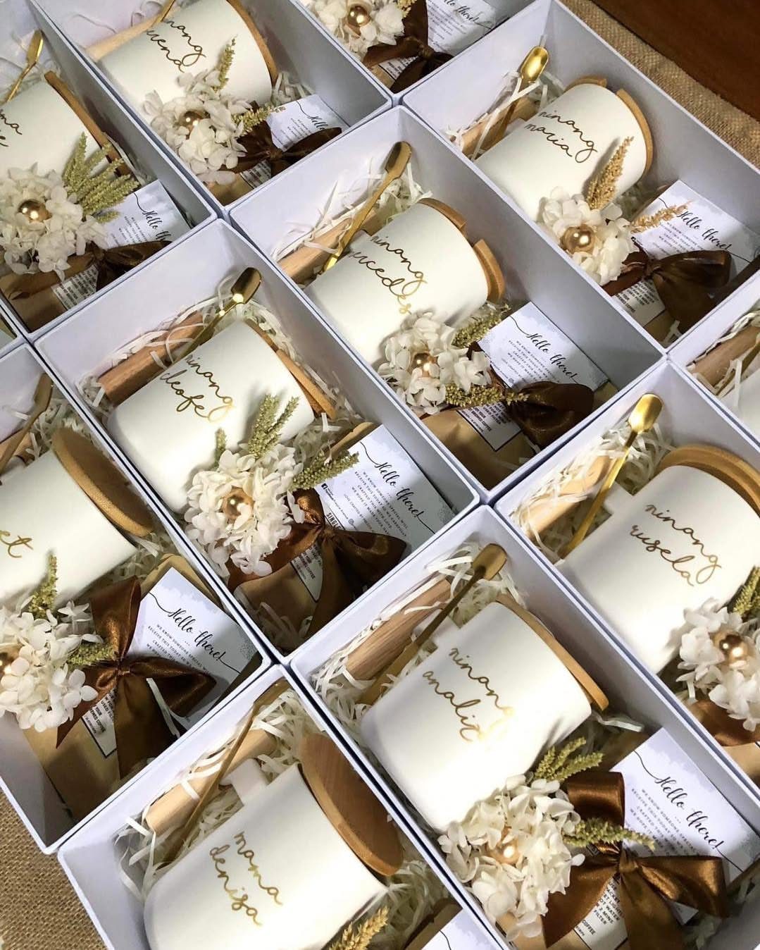 White gift boxes containing ceramic mugs, gold spoons, and white dried flowers.
