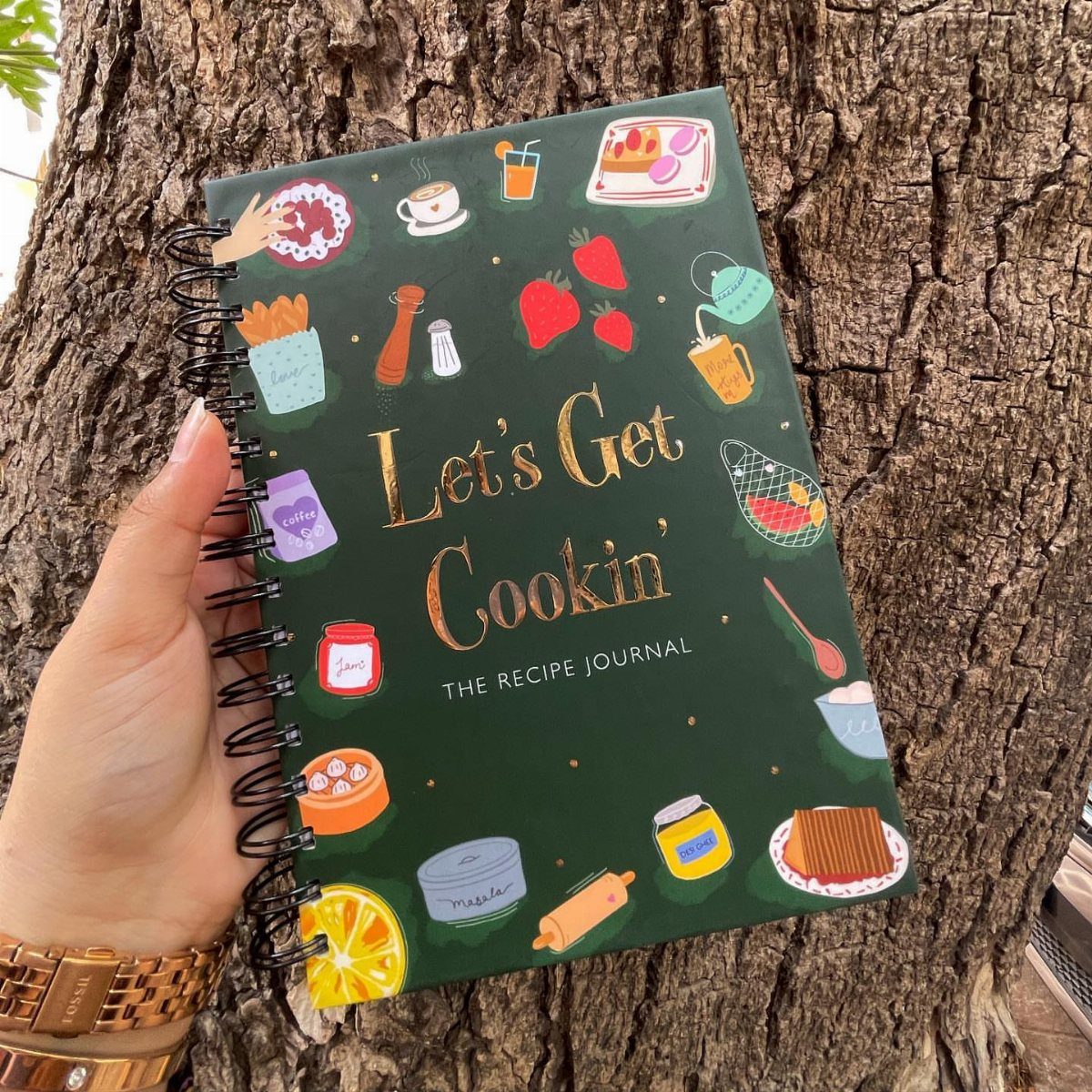 Recipe journal with gold lettering and food illustrations held against tree bark.