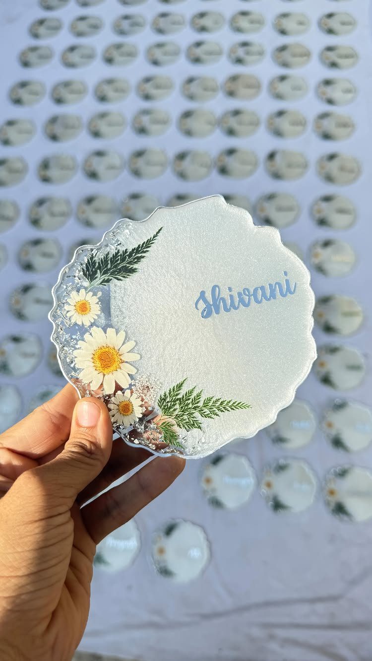 Resin coaster with pressed daisies, ferns, and the name Shivani in blue script.