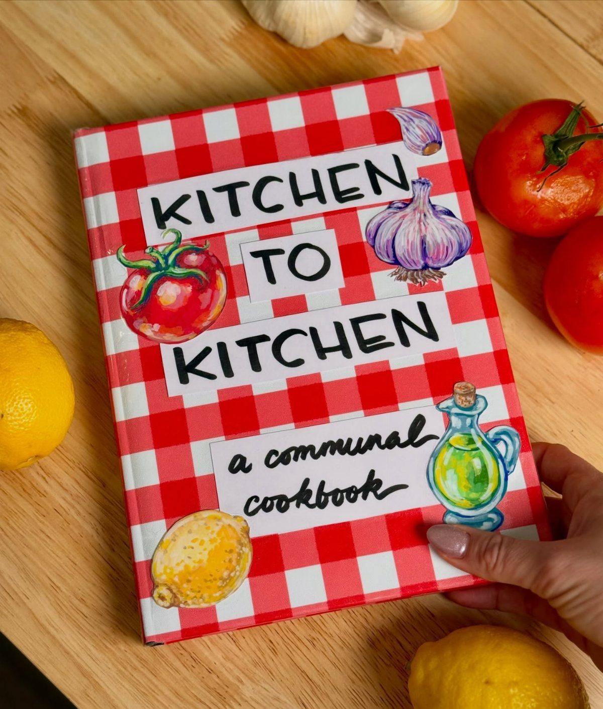 Red and white checkered communal cookbook surrounded by fresh tomatoes, lemons, and garlic.