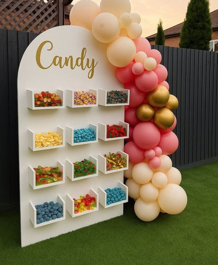 White arched candy wall with colorful sweets and a pink and gold balloon garland.