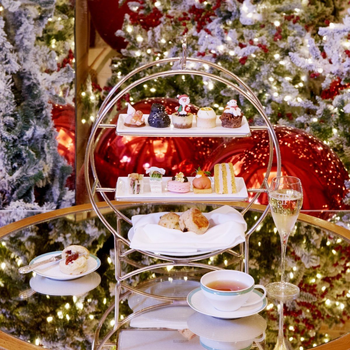 Three-tiered stand with festive pastries, tea, and champagne before a Christmas tree.
