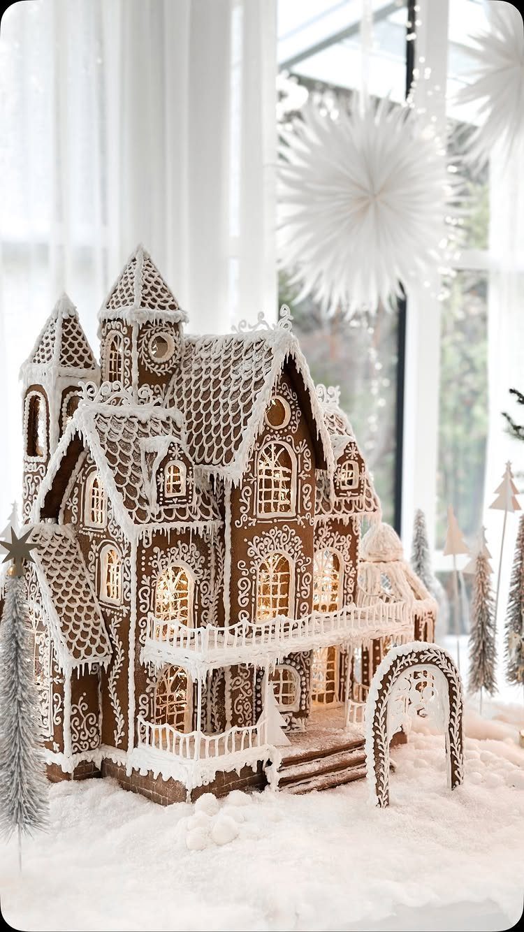Intricate gingerbread house with white icing on artificial snow with frosted trees.