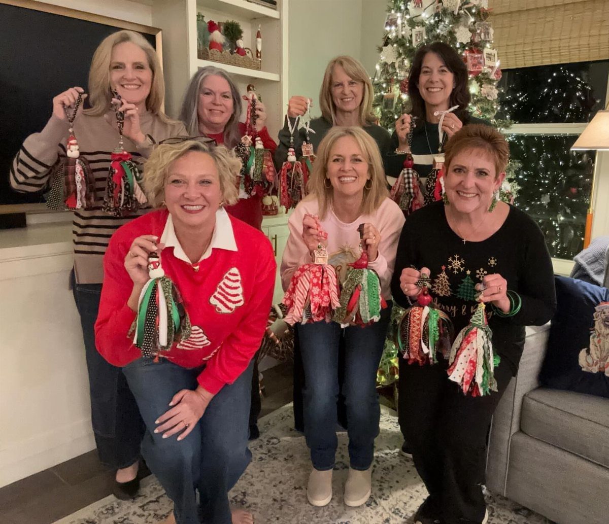 Six women smiling and holding handmade festive fabric tassel ornaments in a living room.