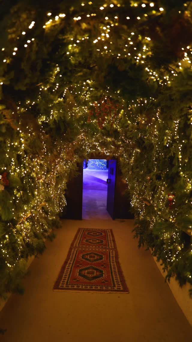 Evergreen branch tunnel with warm fairy lights and a patterned red runner rug.