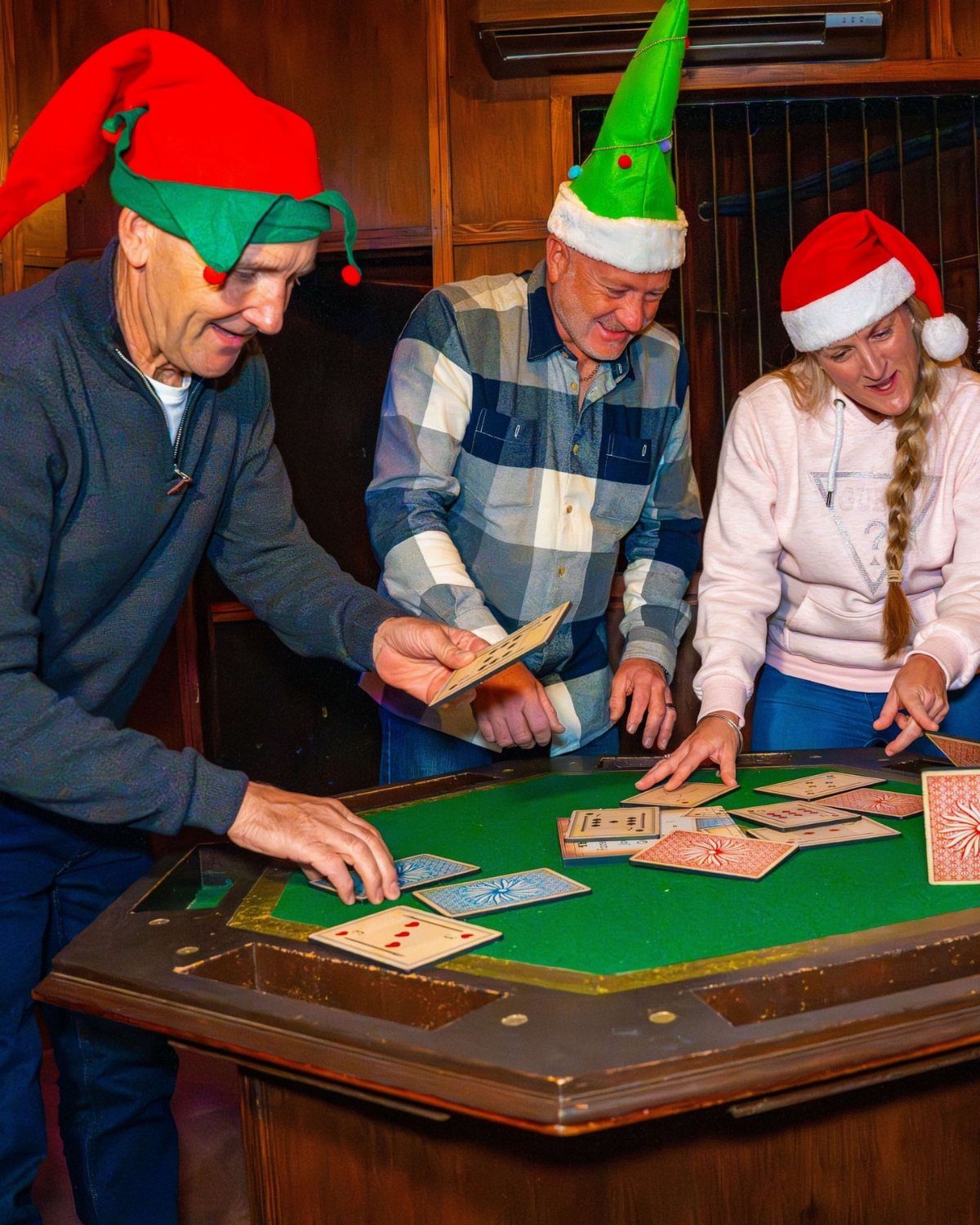 Three people in Christmas hats playing a card game at a green table.