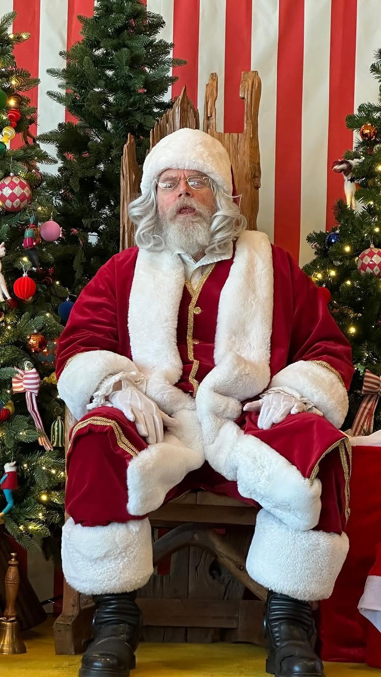 Santa Claus sitting in a wooden throne between two decorated Christmas trees.