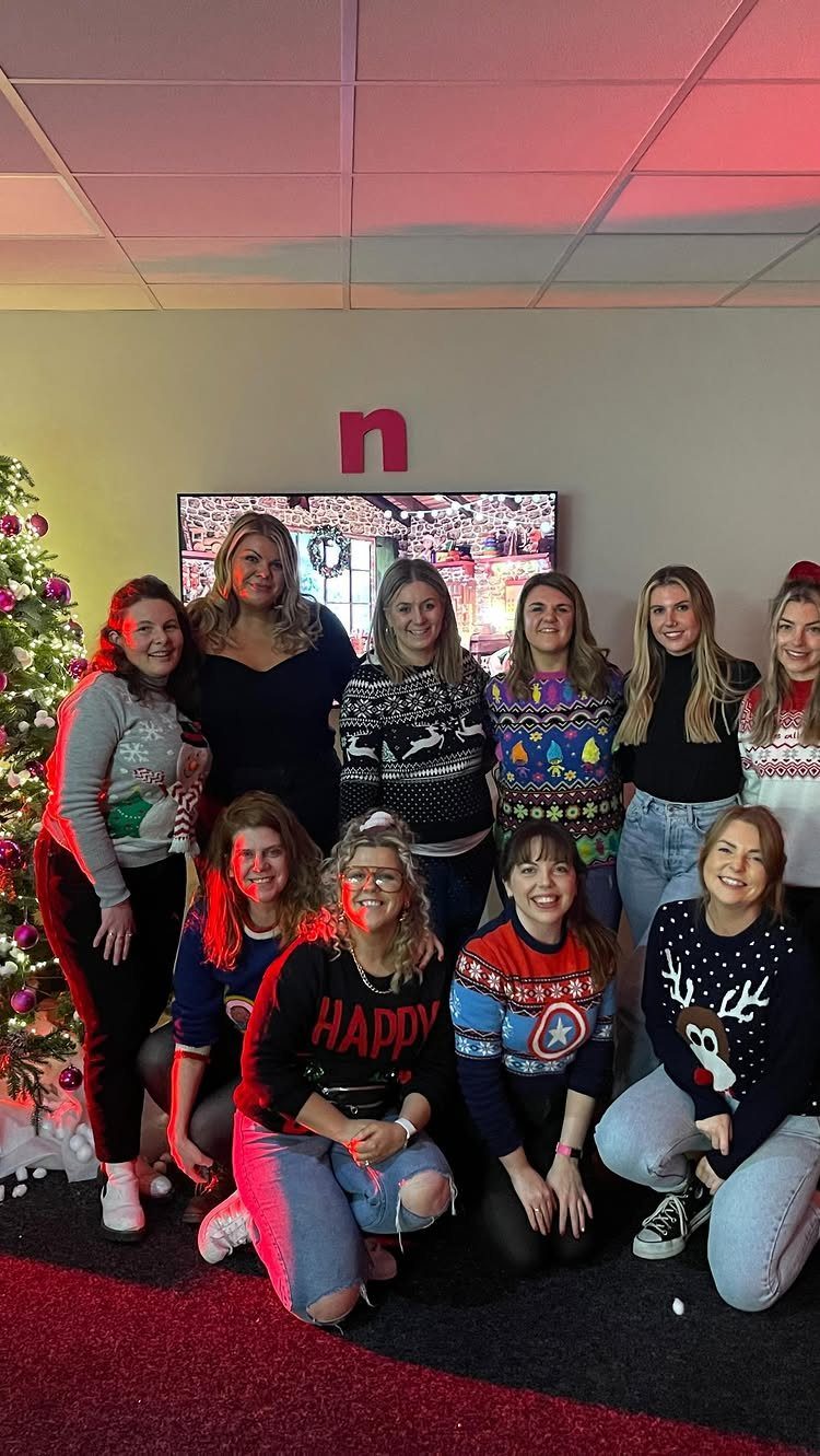 Group of women in festive sweaters posing next to a decorated Christmas tree.