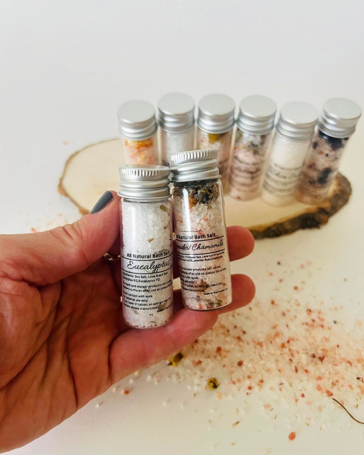 Small glass vials of colorful bath salts held in a hand over wooden slice.