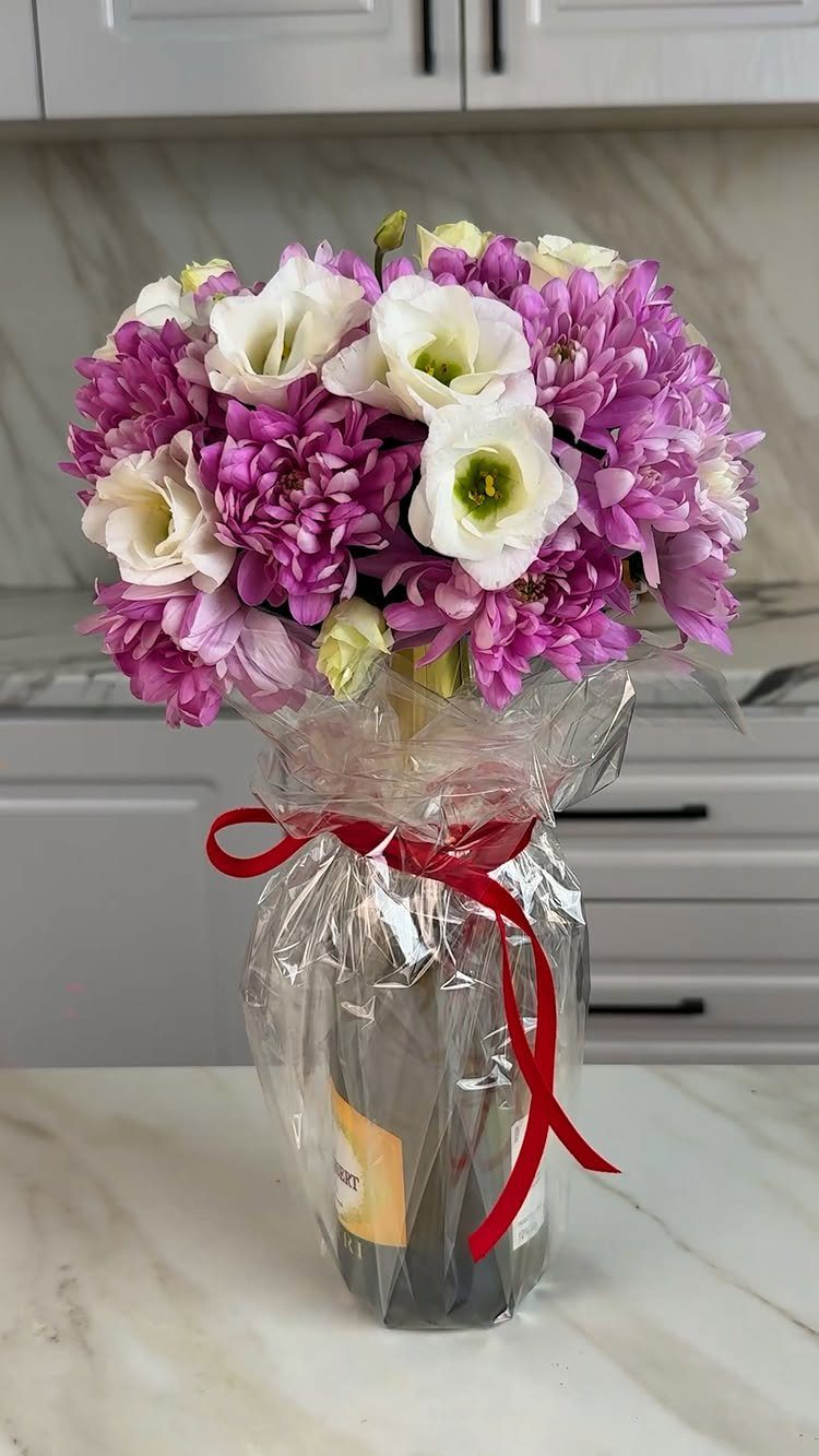 Wine bottle wrapped in cellophane with purple and white flowers and red ribbon.