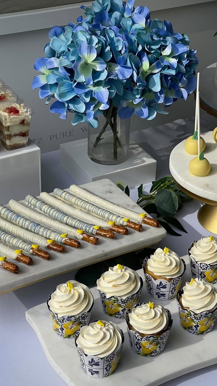 Dessert table with blue hydrangeas, cannoli, and lemon-themed cupcakes on marble slabs.